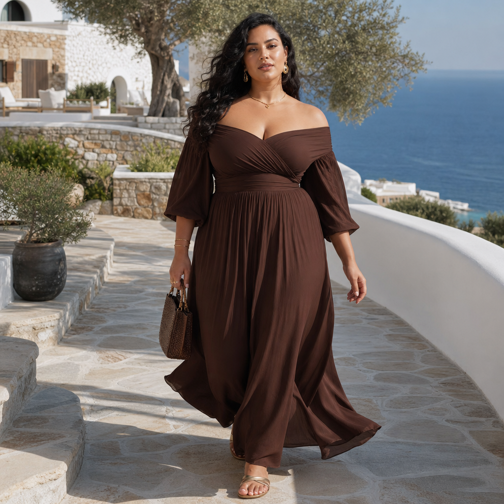 A stylish woman walks along a stone pathway overlooking the ocean, wearing an off-shoulder brown maxi dress with flowing sleeves, gold jewelry, and sandals, carrying a textured handbag against a backdrop of white Mediterranean-style architecture and olive trees.