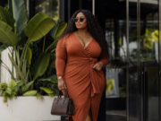 Best Wrap Dresses for Curvy Women: Flattering Fits, Supportive Styles, and How to Choose the Right One A stylish woman in sunglasses and a rust-colored wrap dress poses outside a modern building, carrying a structured handbag beside lush tropical plants.